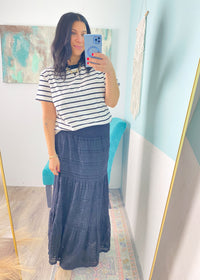 Woman taking a mirror selfie wearing a striped shirt and denim skirt in a room with a colorful painting on the wall.