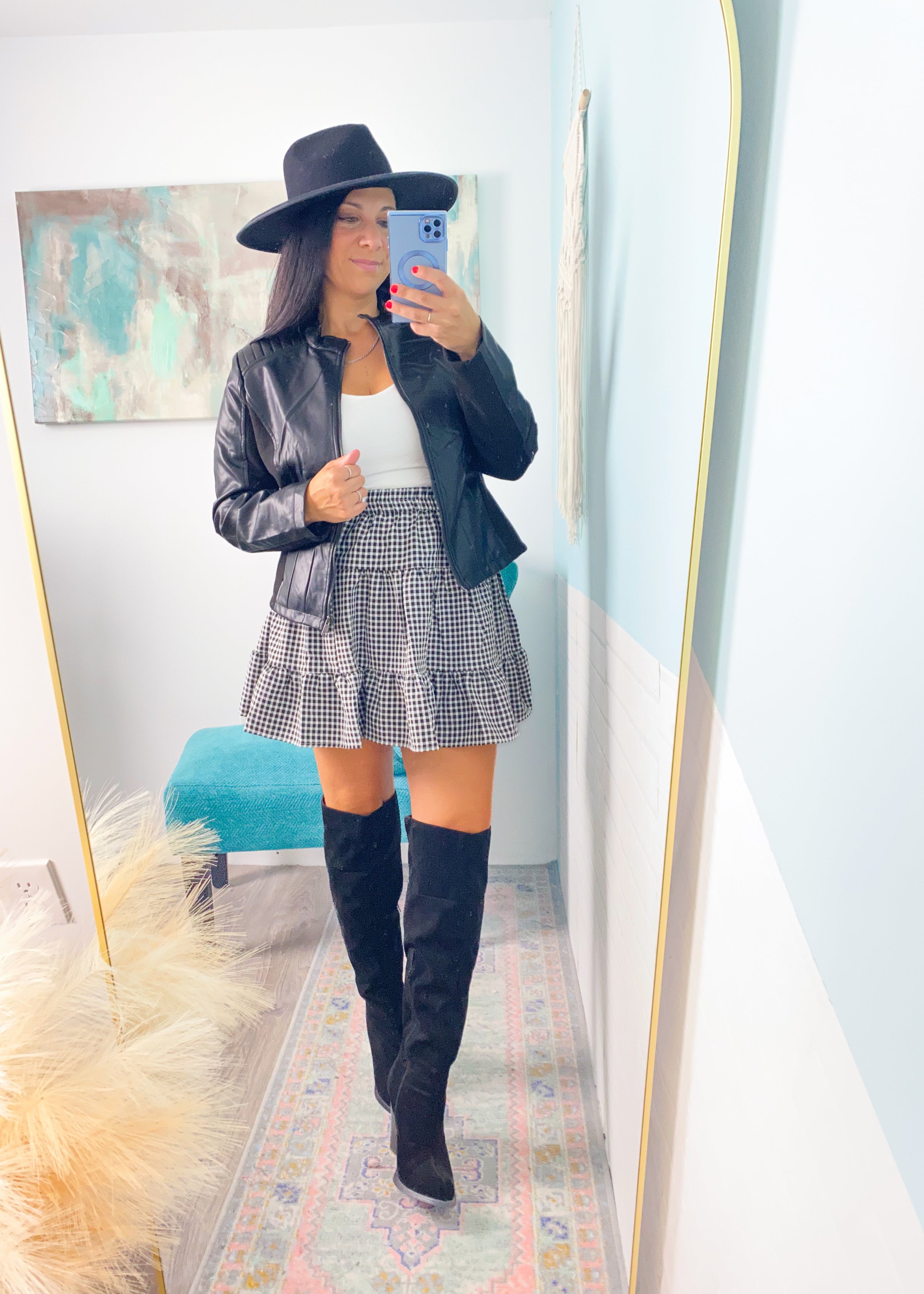 Woman taking a mirror selfie wearing a black leather jacket, white top, plaid skirt, and knee-high boots.