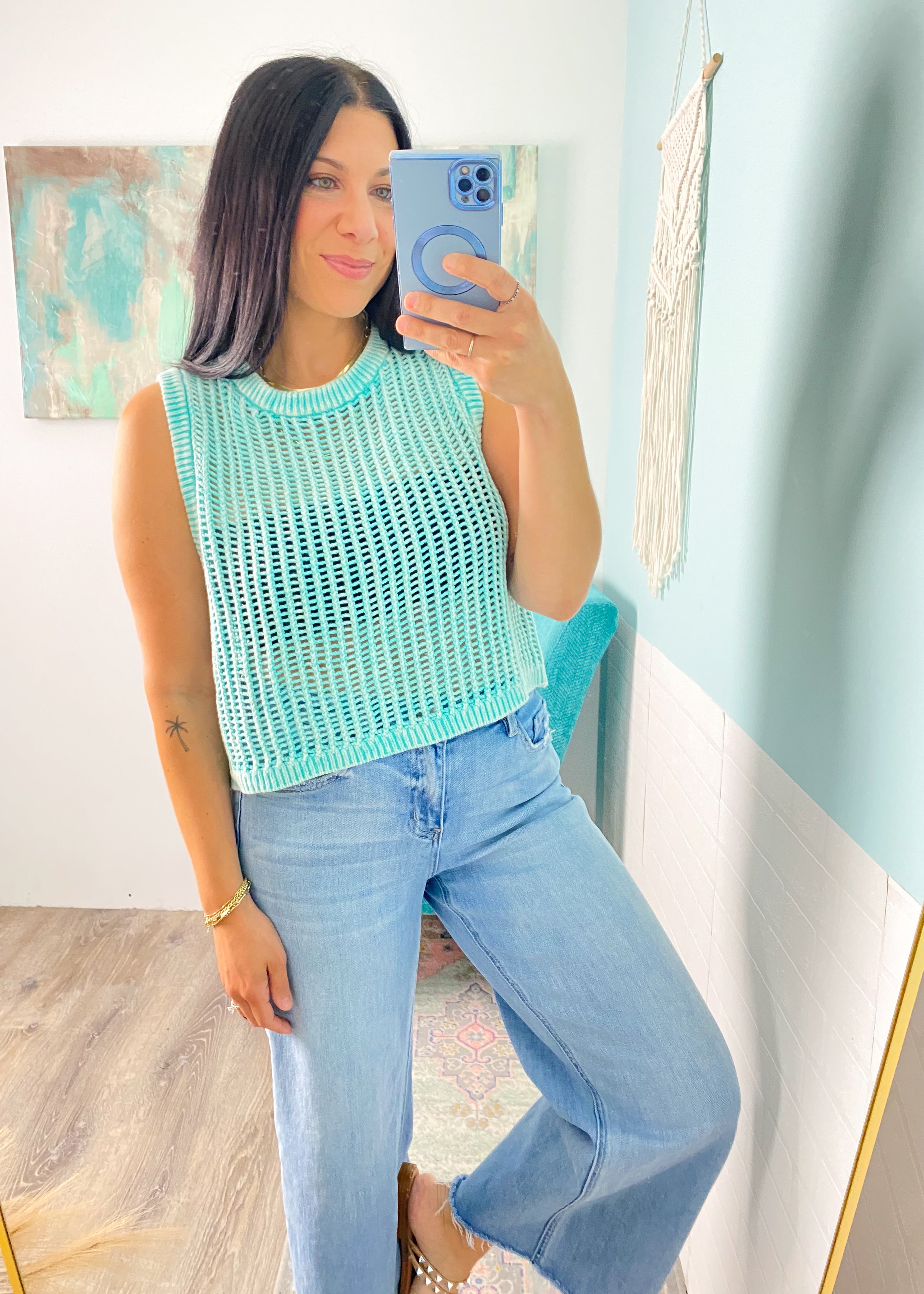Woman taking a mirror selfie wearing a mint green sleeveless crochet top and jeans in a room with light blue walls.