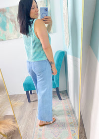 Woman taking a mirror selfie wearing  mint green sleeveless crochet top  and jeans in a room with a colorful rug and teal chair.