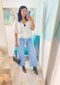 Woman taking a mirror selfie wearing a white lace top, blue jeans, and brown shoes in a room with a colorful rug and wall art.