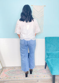 Woman wearing a white eyelet tie front blouse and blue jeans standing in a room with a blue wall and teal chair.