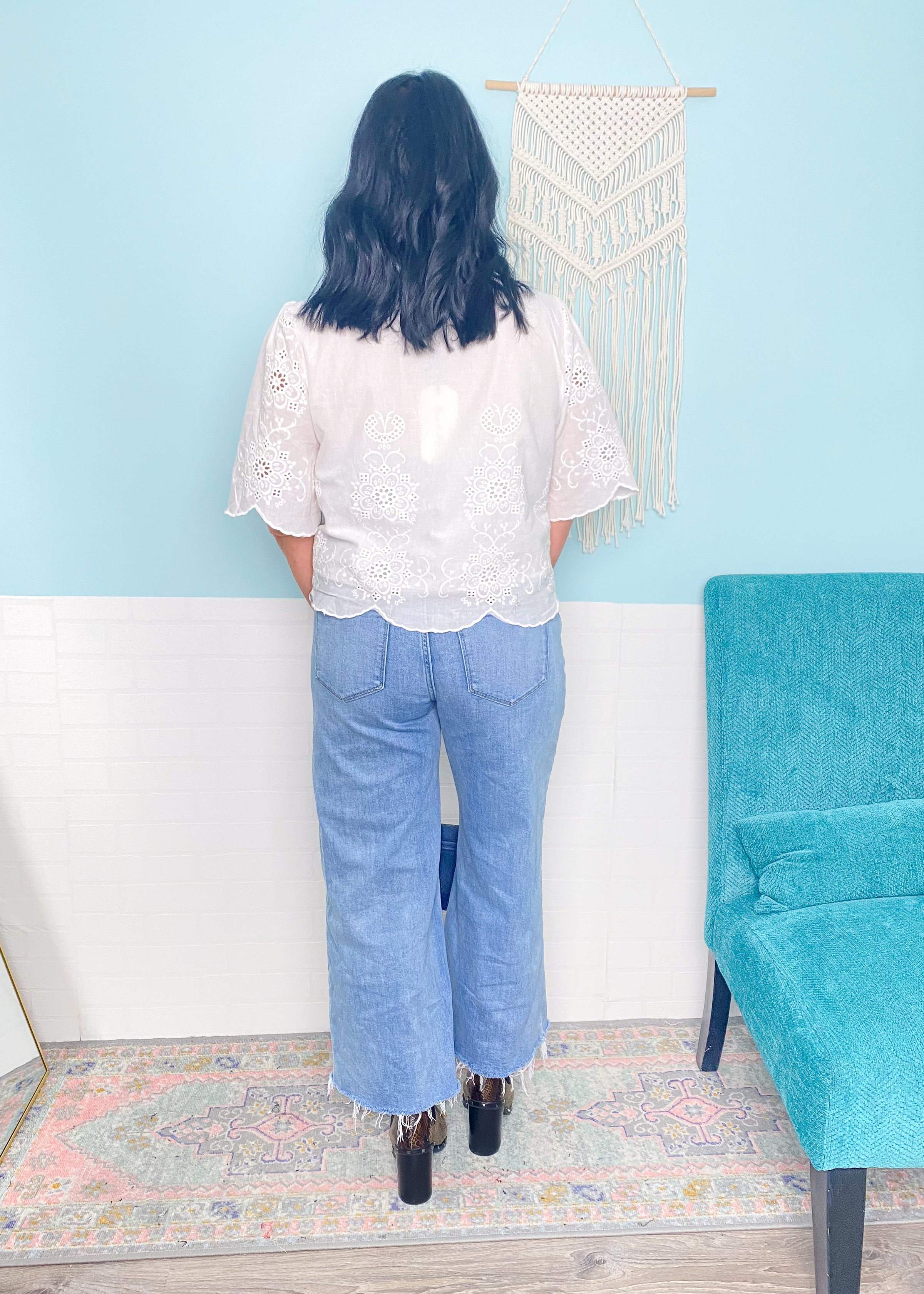 Woman wearing a white eyelet tie front blouse and blue jeans standing in a room with a blue wall and teal chair.