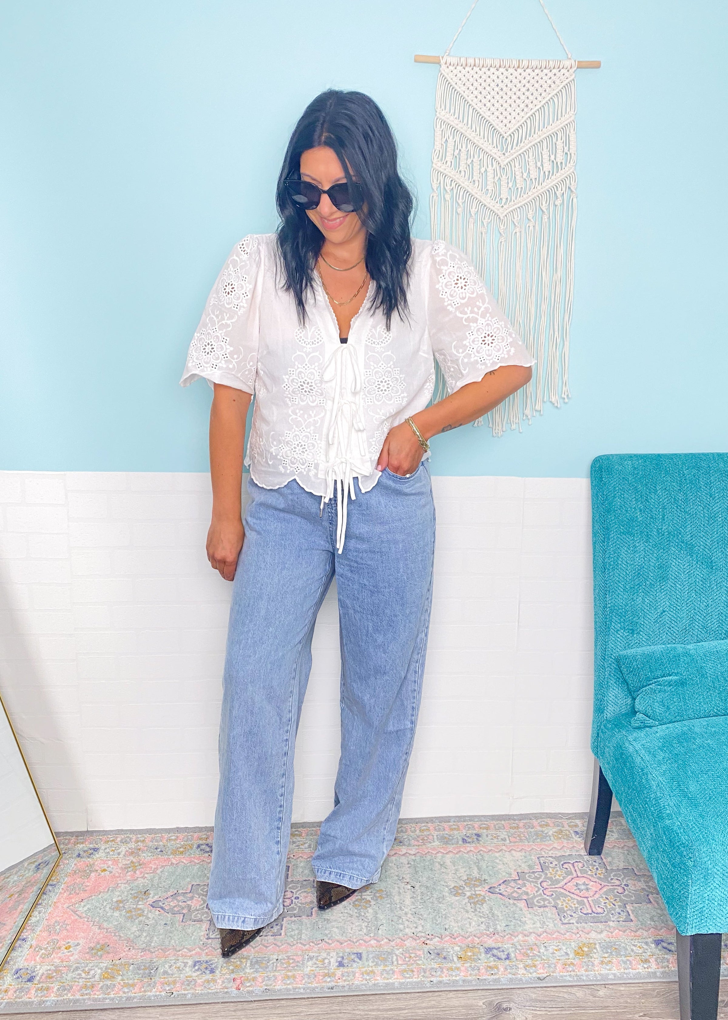 Woman wearing a white eyelet tie front blouse and blue jeans standing in a room with a blue wall and teal chair.