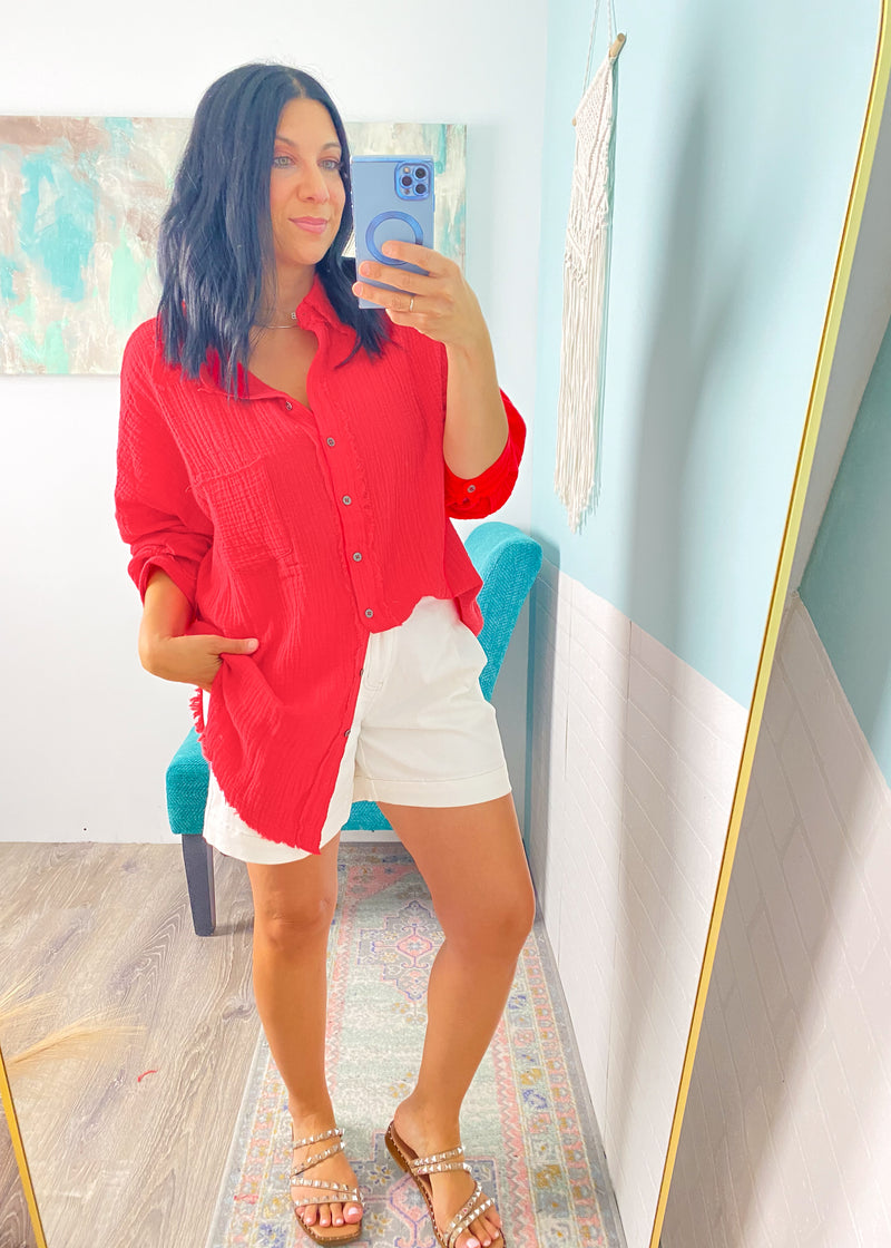 Woman taking a mirror selfie wearing a red shirt and white shorts in a room with a colorful rug and blue chair.