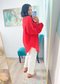 Person wearing a red gauze top and white pants taking a mirror selfie in a room with a colorful rug and abstract painting.