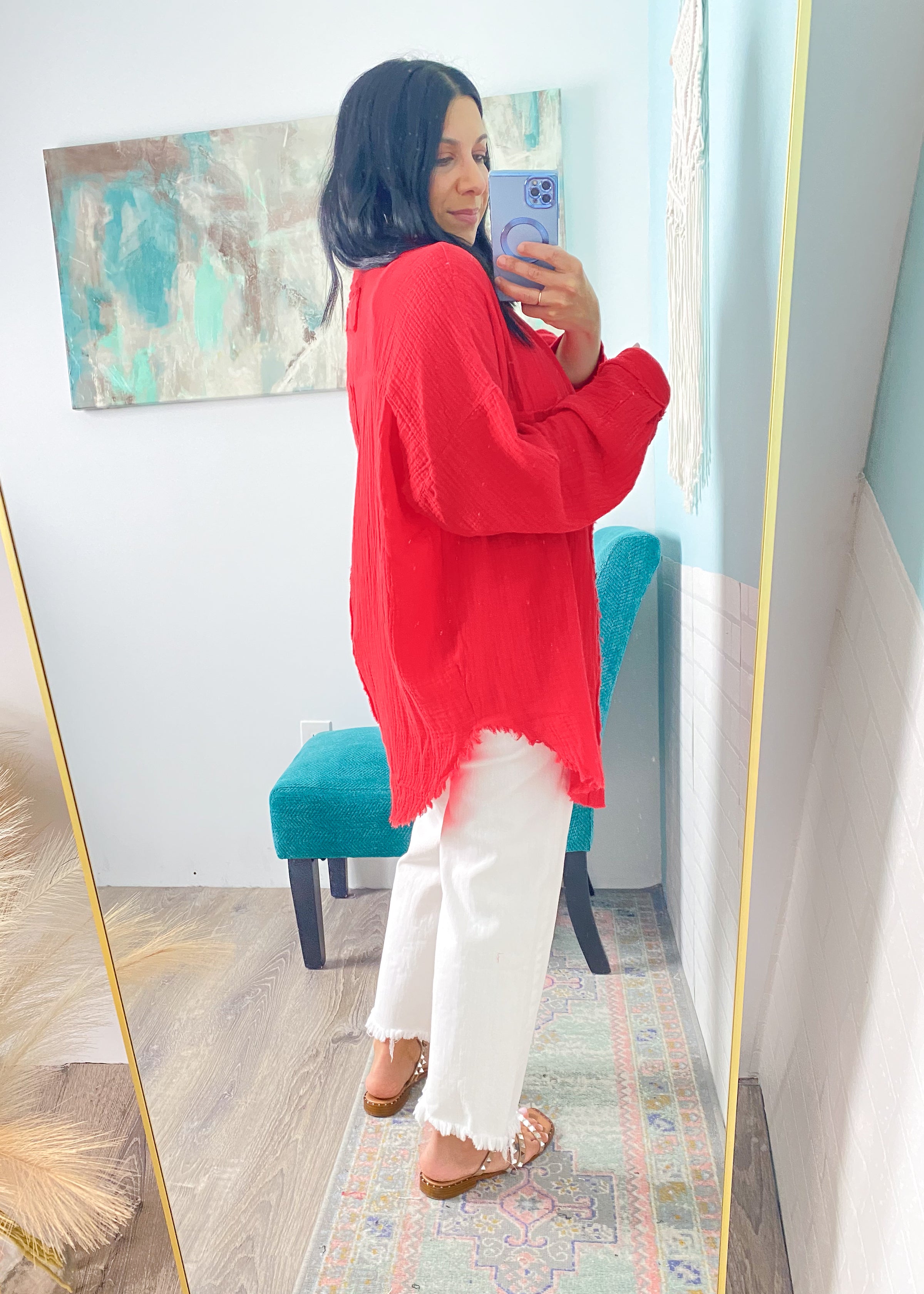Person wearing a red gauze top and white pants taking a mirror selfie in a room with a colorful rug and abstract painting.