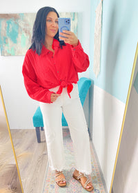 Woman taking a mirror selfie wearing a red shirt and white pants in a room with a colorful rug and abstract painting.