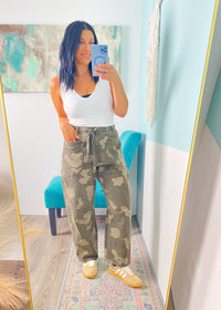 Person taking a mirror selfie wearing a white tank top and camouflage pants in a room with a colorful rug and blue wall.