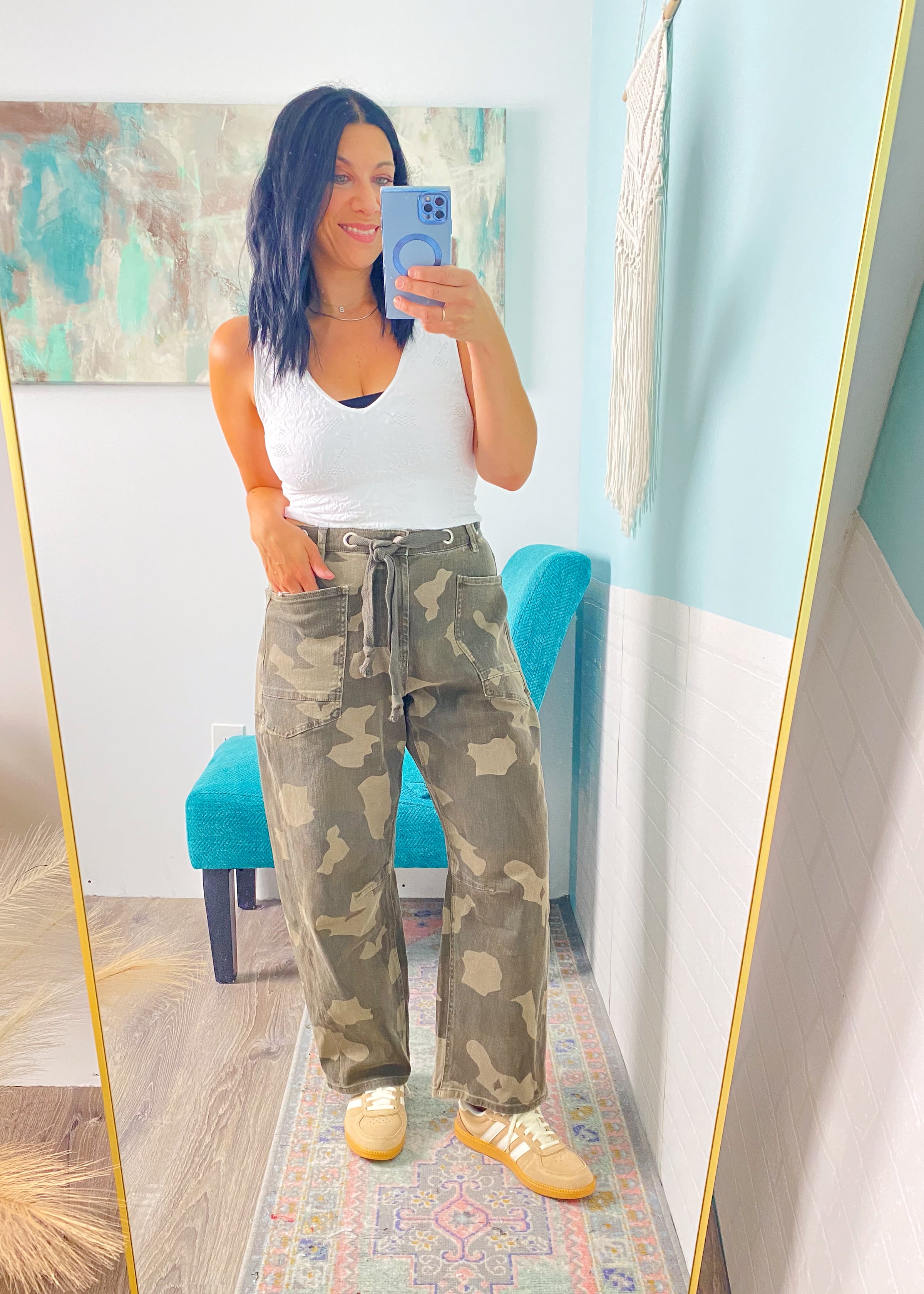 Person taking a mirror selfie wearing a white tank top and camouflage pants in a room with a colorful rug and blue wall.