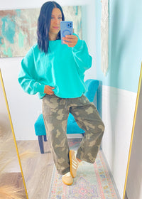 Person wearing a turquoise sweatshirt and camouflage pants taking a mirror selfie.