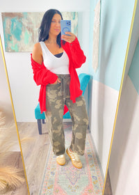 Woman taking a mirror selfie wearing a red cardigan, white top, camouflage pants, and beige sneakers.