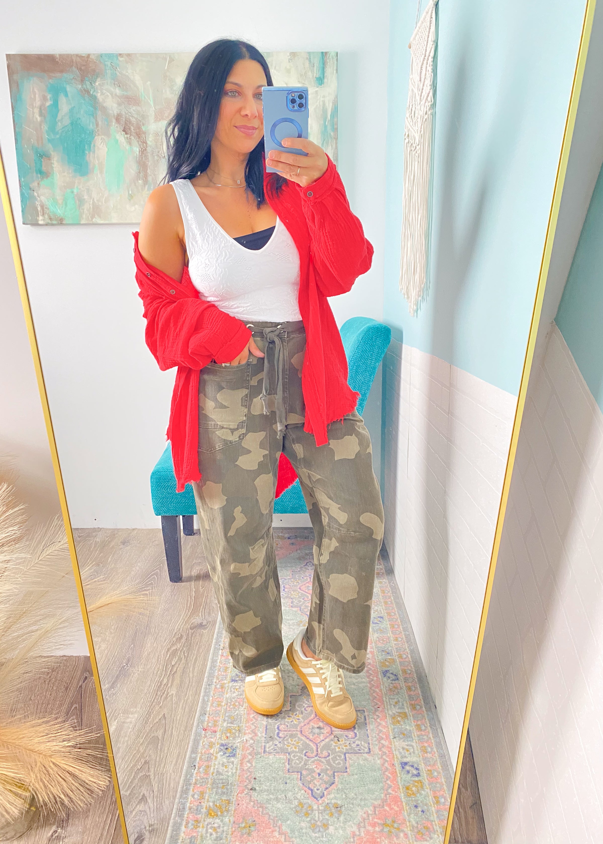 Woman taking a mirror selfie wearing a red top open, white top, camouflage pants, and beige sneakers.