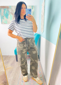 Woman taking a mirror selfie wearing a striped shirt and camouflage pants in a room with a colorful rug.