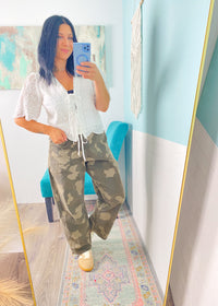 Person taking a mirror selfie wearing a white top and green camouflage pants in a room with light blue walls.
