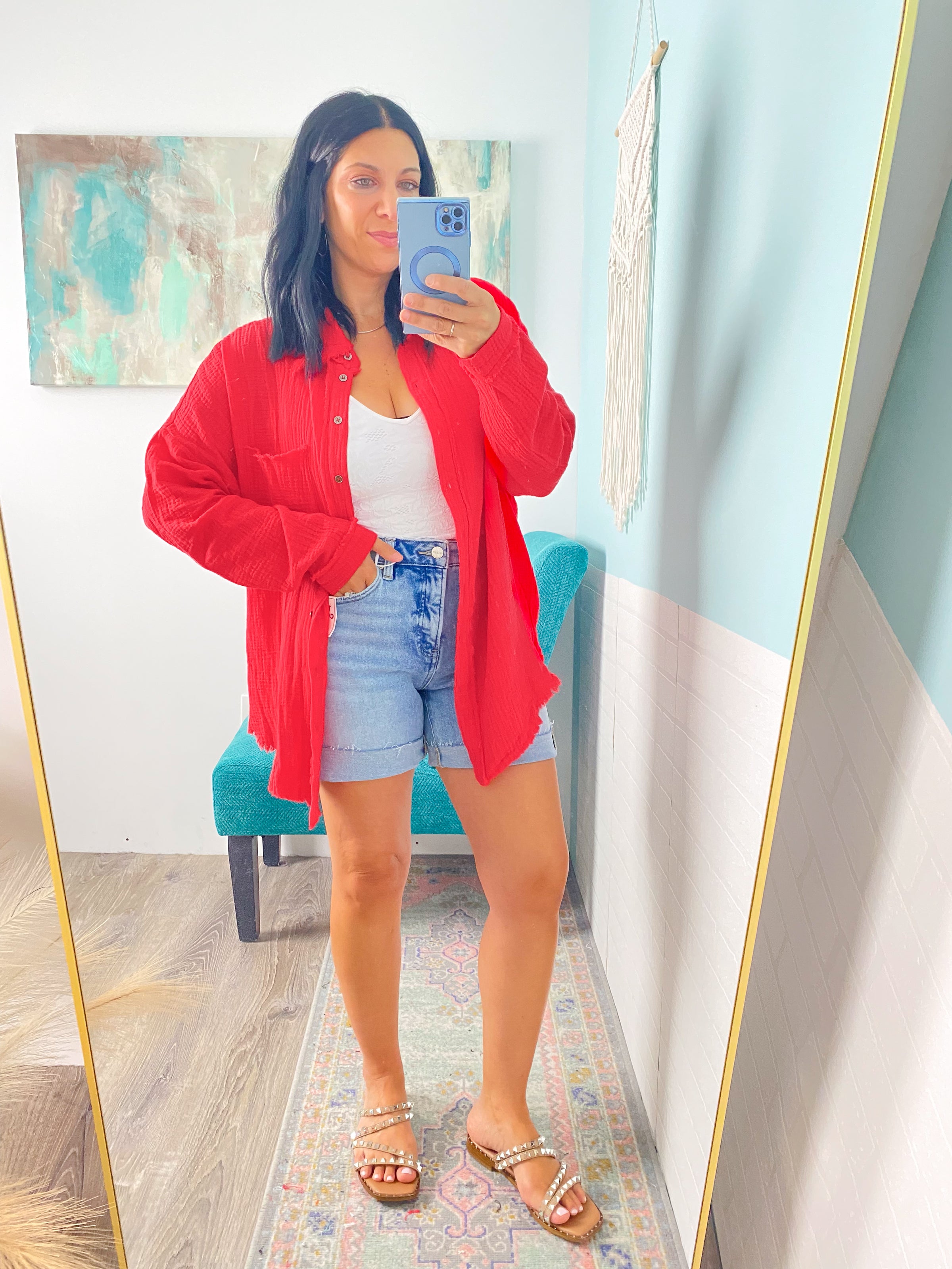 Person wearing a red shirt, white top, and denim shorts taking a mirror selfie.