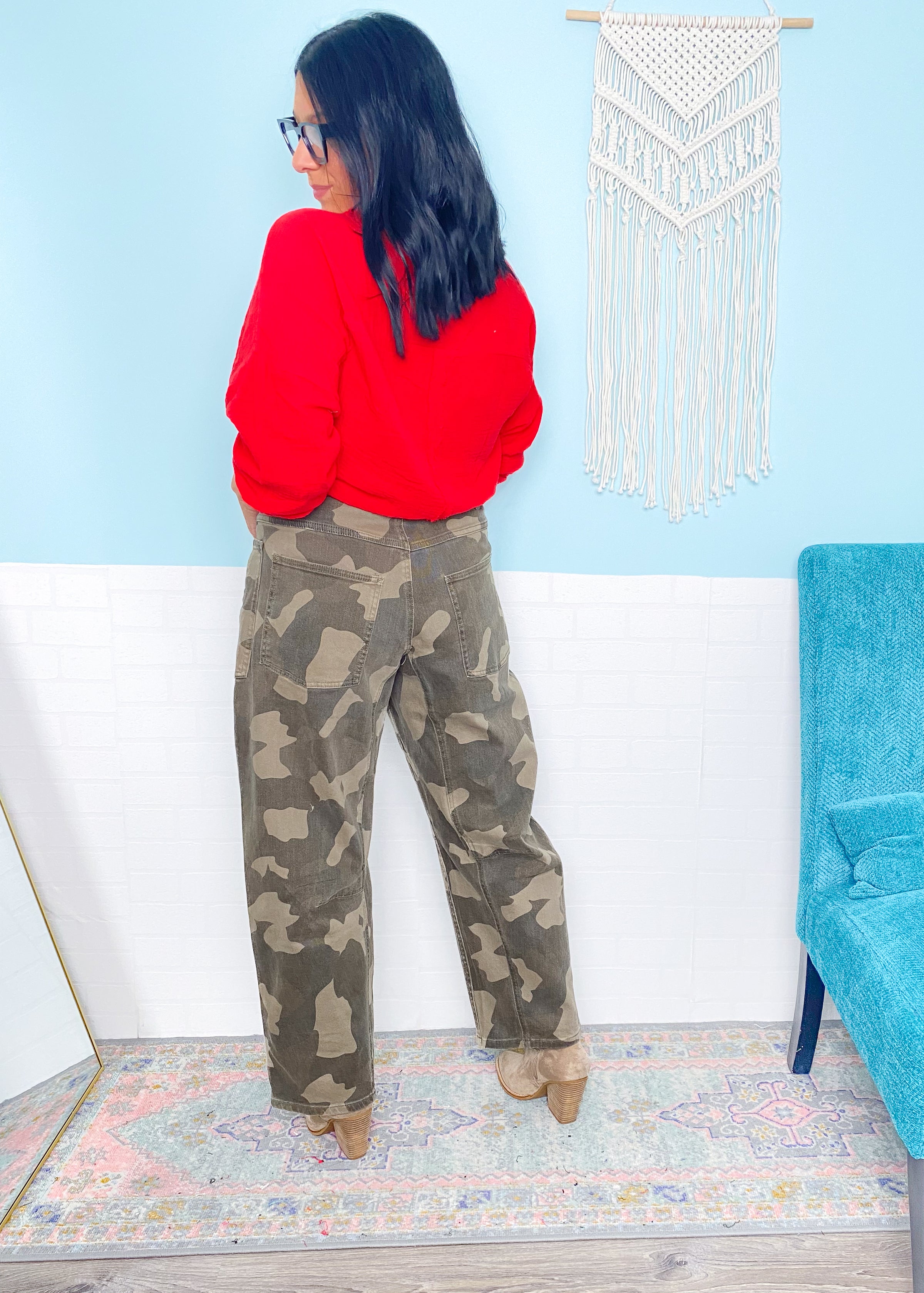 Person wearing a red sweater and camouflage pants in a room with a blue chair and decorative wall hanging.