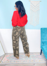 Woman wearing a red top and camouflage pants standing in a room with a blue chair and decorative wall hanging.