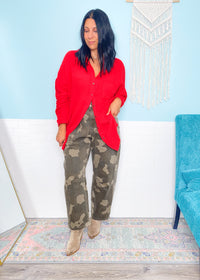 Woman wearing a red shirt and camouflage pants standing in a room with a blue chair and decorative wall hanging.