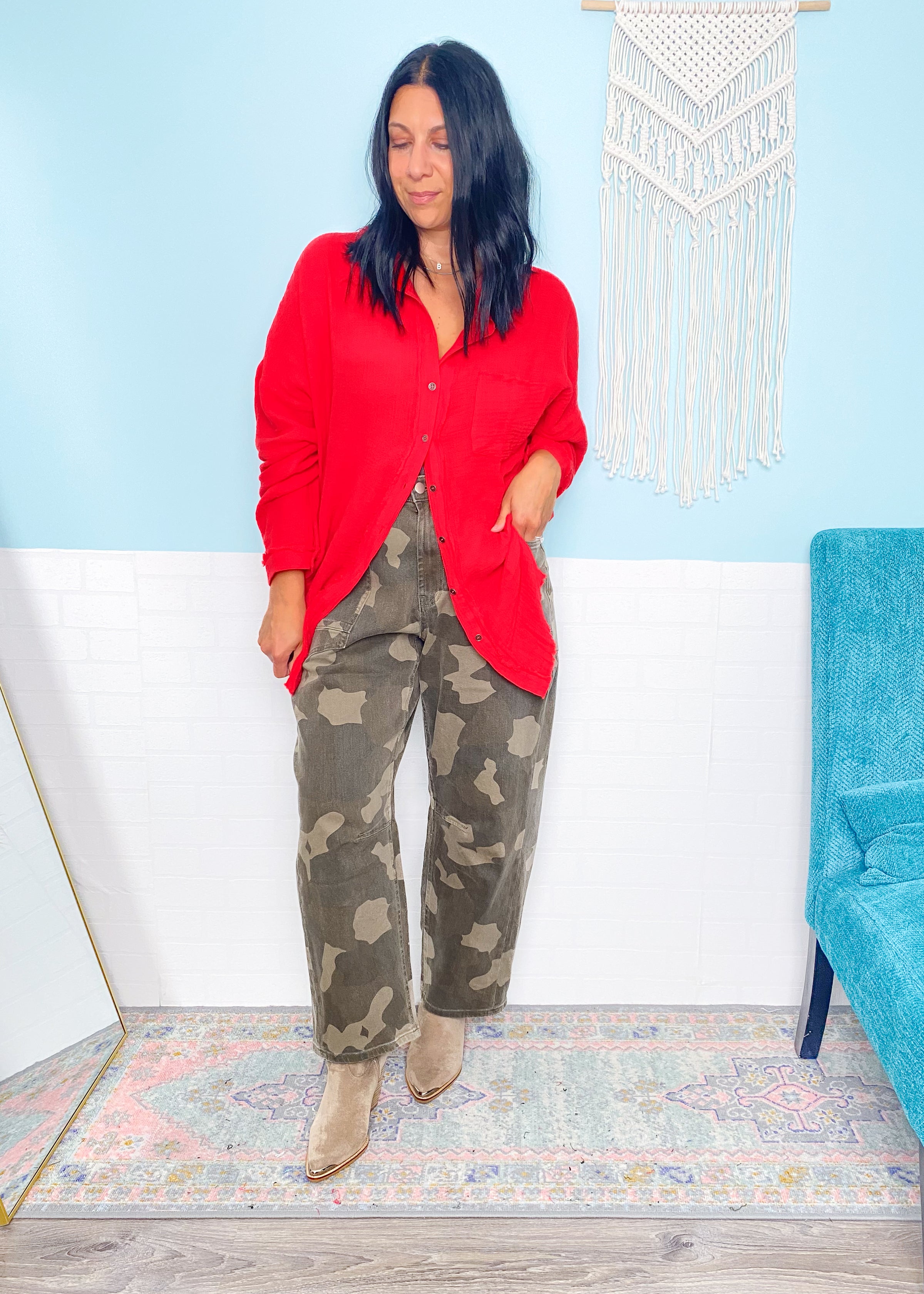 Woman wearing a red shirt and camouflage pants standing in a room with a blue chair and decorative wall hanging.