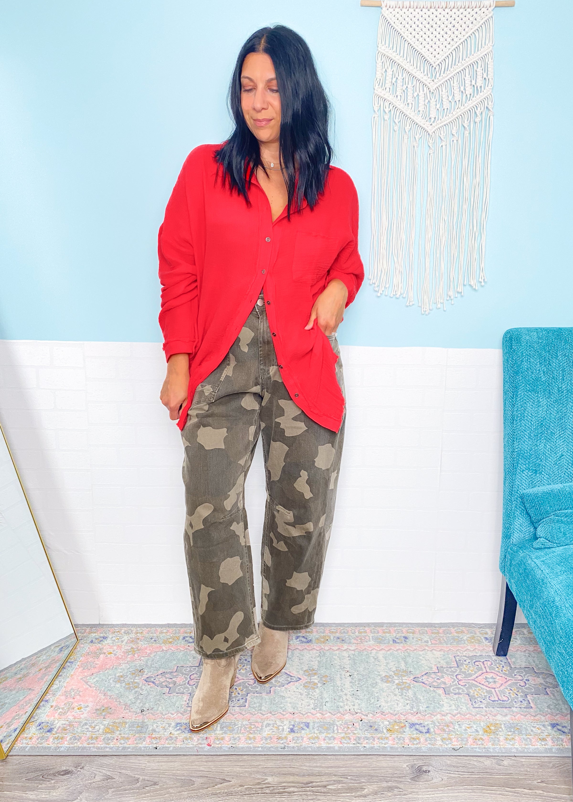 Woman wearing a red shirt and camouflage pants standing in a room with a blue chair and decorative wall hanging.